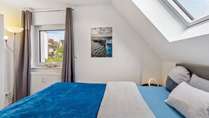 A bedroom with a skylight, a bed with blue and white bedding, a nightstand with a lamp, and a painting of a coastal scene. - Modern Apartment – Quiet & Prime Location in Neheim (Arnsberg)