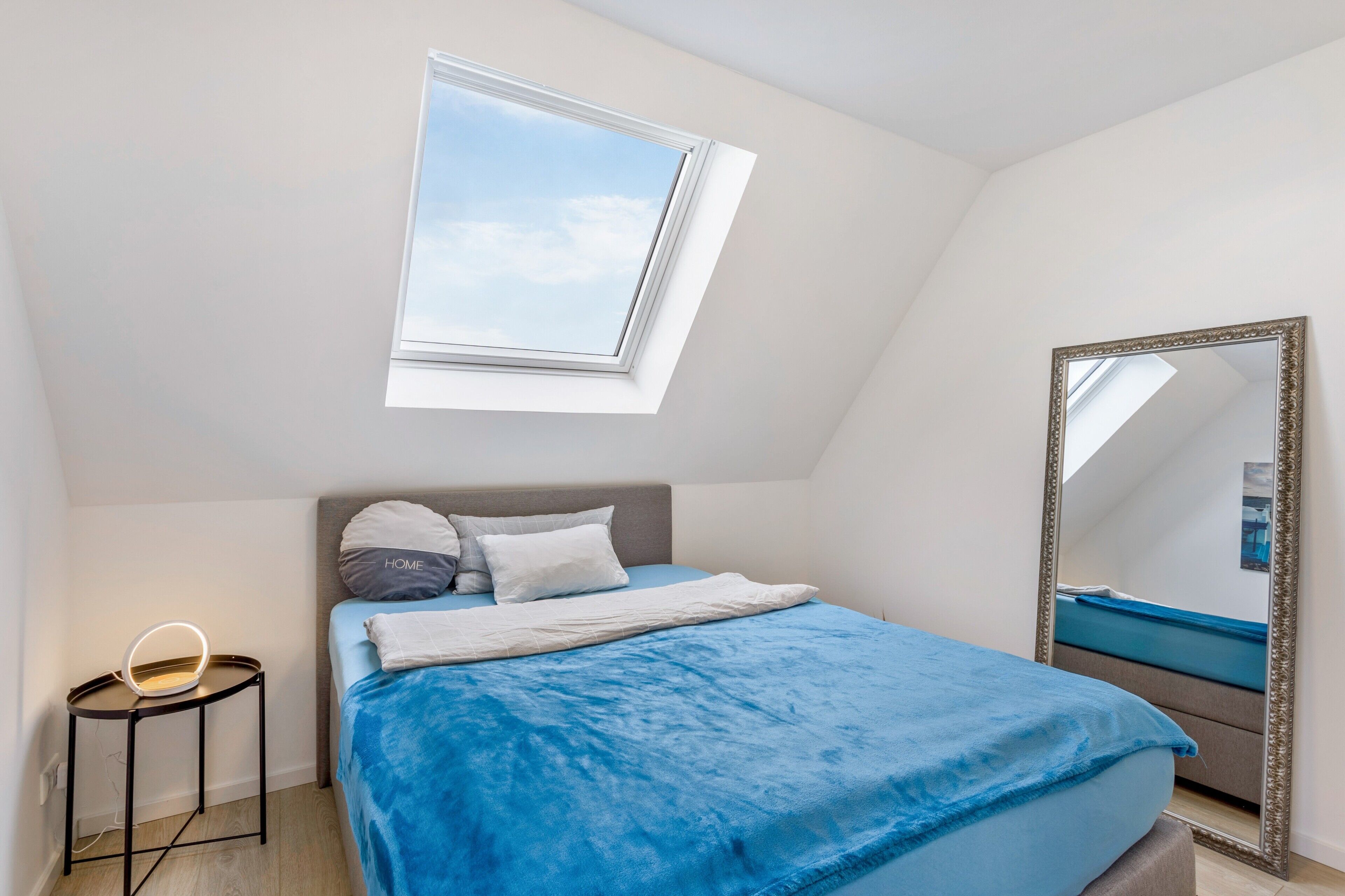 A bedroom with a skylight, a bed with a blue cover, a bedside table with a lamp, and a large mirror.