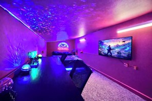 Una sala de juegos con una mesa larga, sillas, un letrero de neón, un televisor grande y una lámpara en el techo.