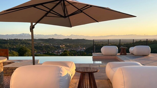 An outdoor seating area with a pool, white cushioned chairs, a wooden table, and a large umbrella. - Junior Suite with SPA & Pool (Dogliani)