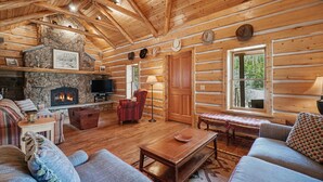A log cabin interior with a stone fireplace, wooden walls, a sofa, a bench, and a television. - Historical Crocker Ranch - Rowe House 23-zone3423 3 Bedroom Home (Drake)