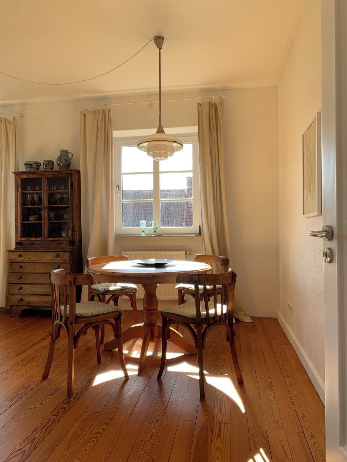 Ein Esszimmer mit einem runden Tisch, Holzstühlen, einem Schrank und einem Fenster mit Vorhängen.