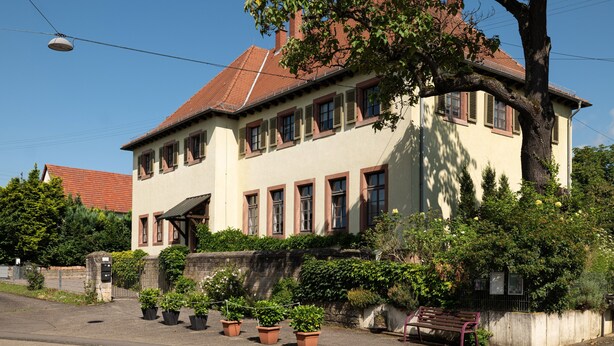 Ein zweistöckiges Gebäude mit rotem Ziegeldach, zahlreichen Fenstern und einem großen Baum davor.