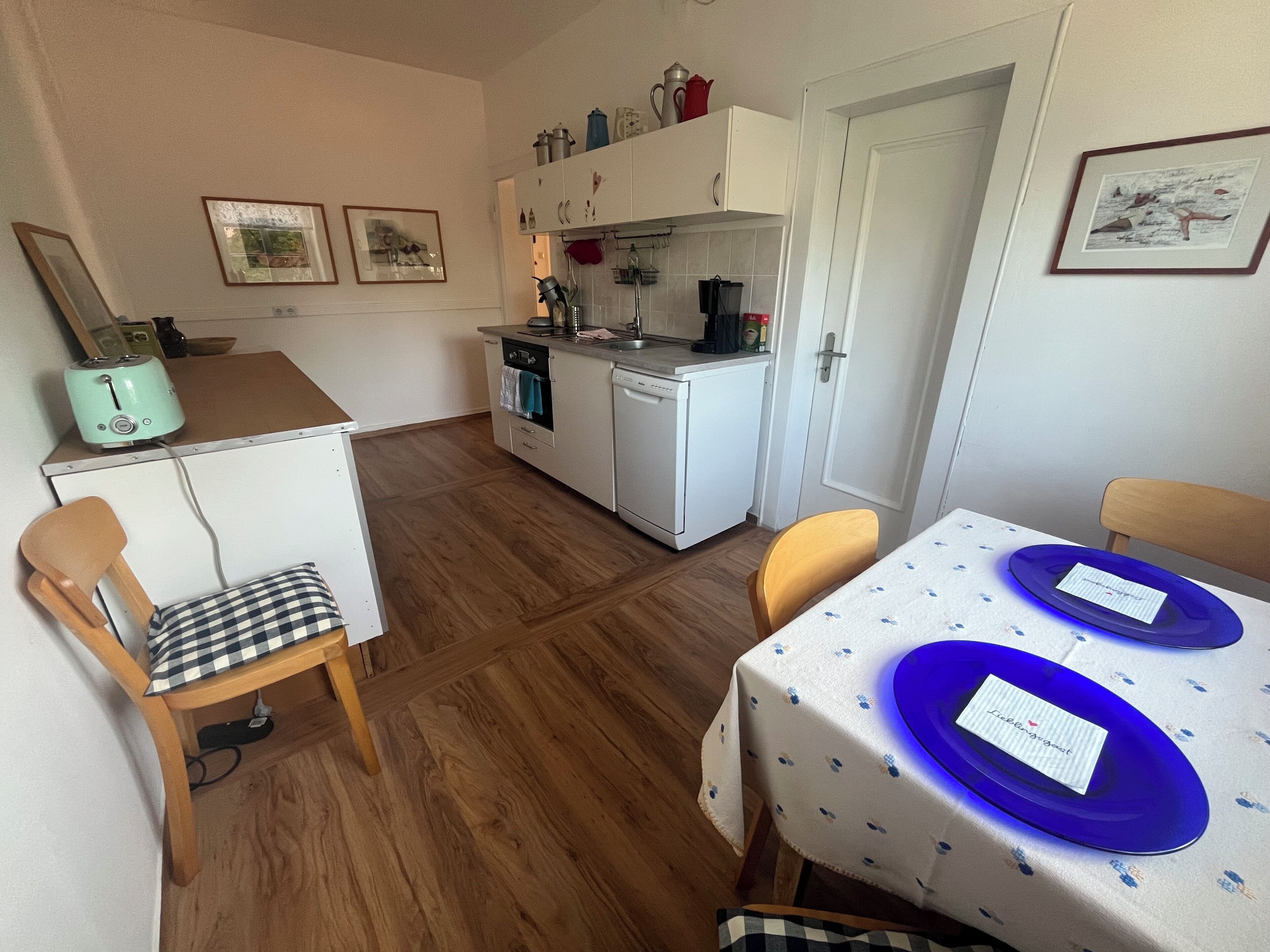 A compact kitchen with a toaster, oven, and sink. There are two chairs and a table with blue plates.