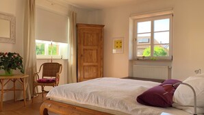 Ein Schlafzimmer mit einem hölzernen Bett, einem Sessel, einem Tisch, einem Fenster mit Aussicht und einer Heizung.