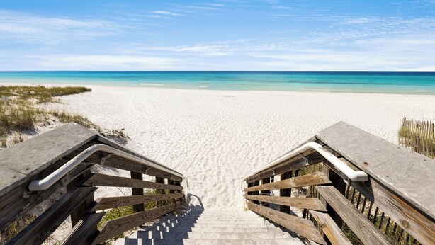 Eine Holztreppe führt zu einem Sandstrand unter blauem Himmel.