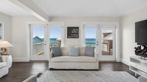 Ein Wohnzimmer mit einem weißen Sofa, einem Teppich und einer TV-Konsole. Zwei Türen führen zu einem Balkon mit Blick aufs Meer und einer Holzterrasse. Ein Schild an der Wand trägt die Aufschrift 'slow down to enjoy the journey'.