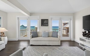 A living room with a white sofa, a rug, and a TV stand. There are two doors leading to a balcony with a view of the ocean and a wooden deck. A sign on the wall reads 'slow down to enjoy the journey.' - Sophisticated 5 BR, Bikes, & Private Beach Access + Under New Management (Santa Rosa Beach)