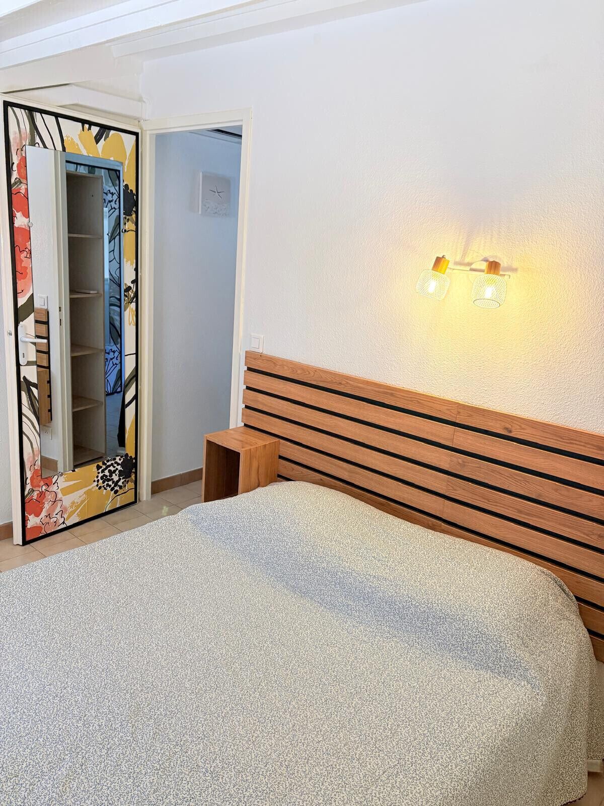 Une chambre à coucher comprenant un lit, une tête de lit en bois, une table de chevet, une armoire avec un miroir et un luminaire fixé au mur.
