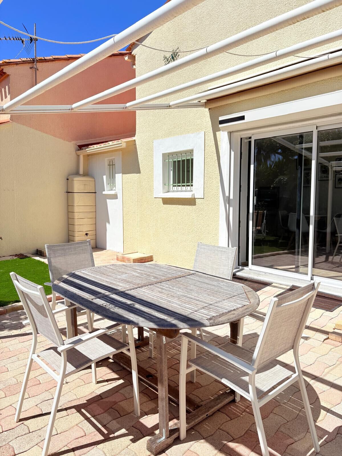 Um pátio externo com mesa e cadeiras, um reservatório de água e uma porta de vidro deslizante que dá para uma área coberta.