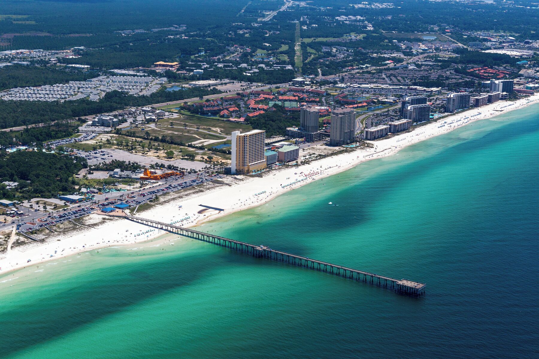 Vista aérea de una ciudad costera con un muelle largo que se adentra en el mar, rodeada de una playa, hoteles y una zona urbana densa.