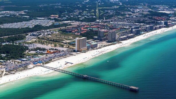 Una vista aerea di una città costiera con un lungo molo che si protende verso il mare, circondata da una spiaggia, hotel e una zona urbana densa.