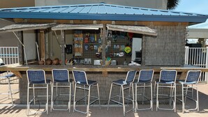 Un piccolo bar all'aperto con tetto blu, un bancone in legno e sei sgabelli da bar in metallo.