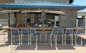 Eine kleine Bar im Freien mit blauem Dach, einer Holztresen und sechs Metallbarhocker.