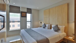 Una habitación de hotel moderna con una cama grande, cabecera de madera, mesita de noche y televisor montado en la pared, además de un ventanal con vistas a edificios.