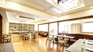A spacious dining area with wooden flooring, multiple tables, and chairs. There are shelves with items on the left wall, and a large window on the right.