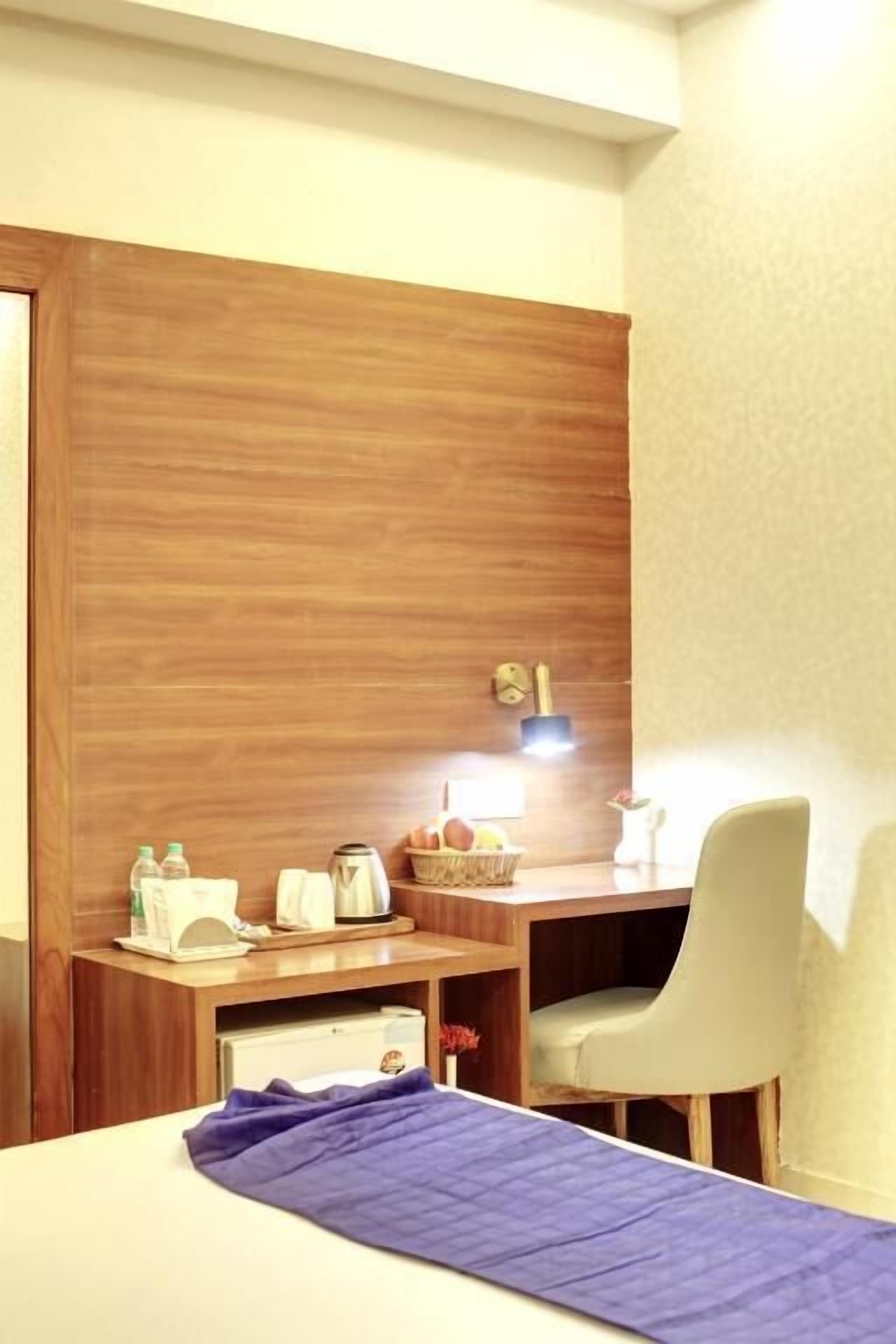 Ein Hotelzimmer mit einem Holztisch, einem Sessel, einem Bett mit blauer Decke, einem kleinen Tisch mit Pflegeprodukten und einer Wanduhr.