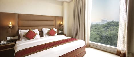 Habitación de hotel con una cama grande, cabecera, lámparas de noche, mesitas de noche y vista a un edificio a través de una ventana.