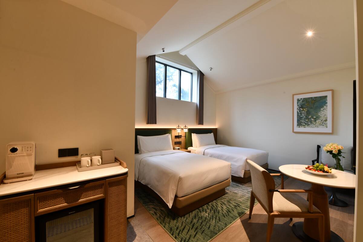 Habitación de hotel con dos camas, un escritorio con cafetera, una mesa de comedor con un florero y un ventanal con cortinas.
