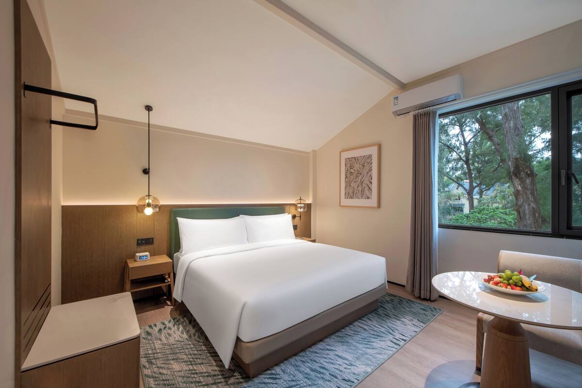 Habitación de hotel con cama, mesita de noche, lámpara, ventana con cortinas y una mesa de comedor con fruta.