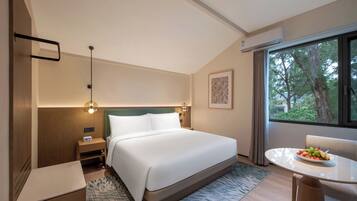 Habitación de hotel con cama, mesita de noche, lámpara, ventana con cortinas y una mesa de comedor con fruta.