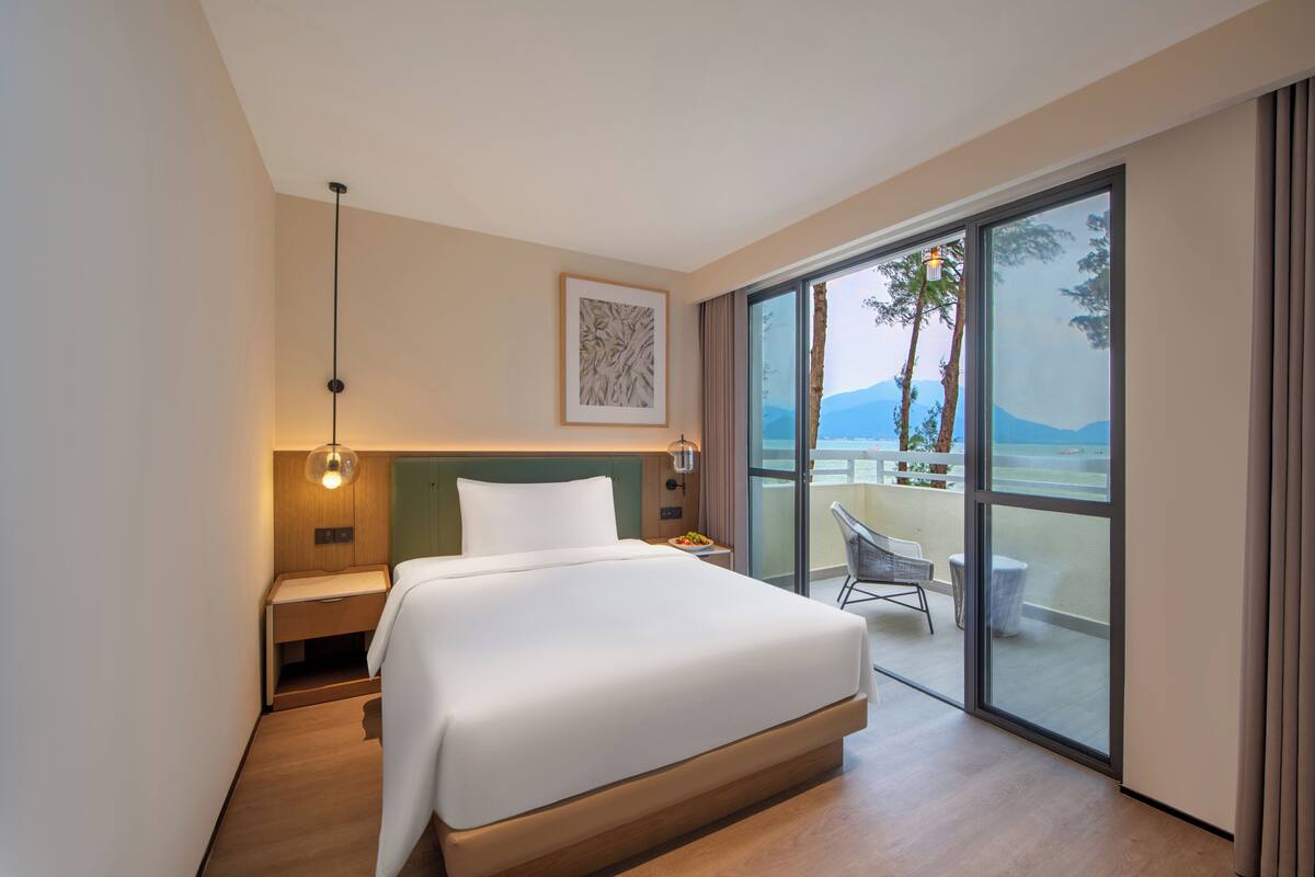Una habitación de hotel moderna con una cama grande, una mesita de noche, un cuadro en la pared y un balcón con una silla.