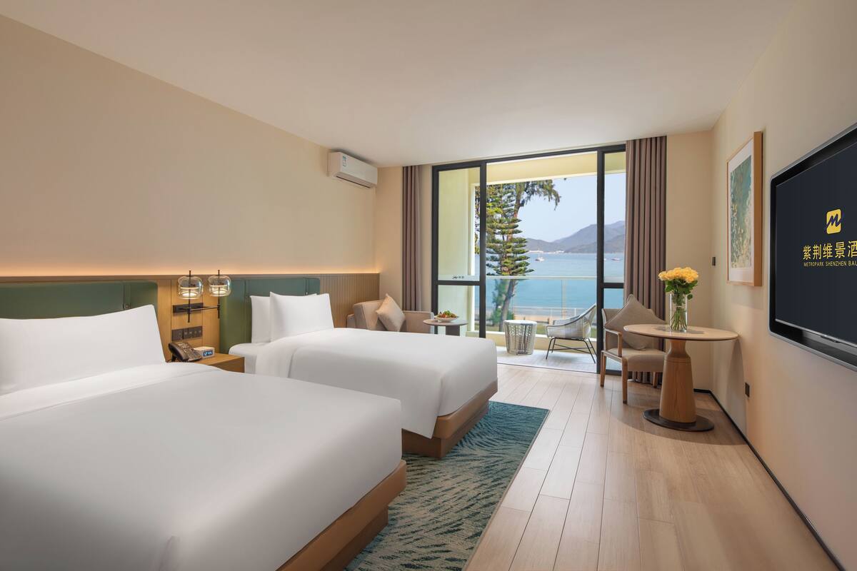 Habitación de hotel con dos camas, un televisor, una mesita con un florero y un balcón con vistas al mar.