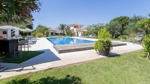 Piscina com deck ladrilhado, rodeada por vegetação e um edifício de dois andares ao fundo.