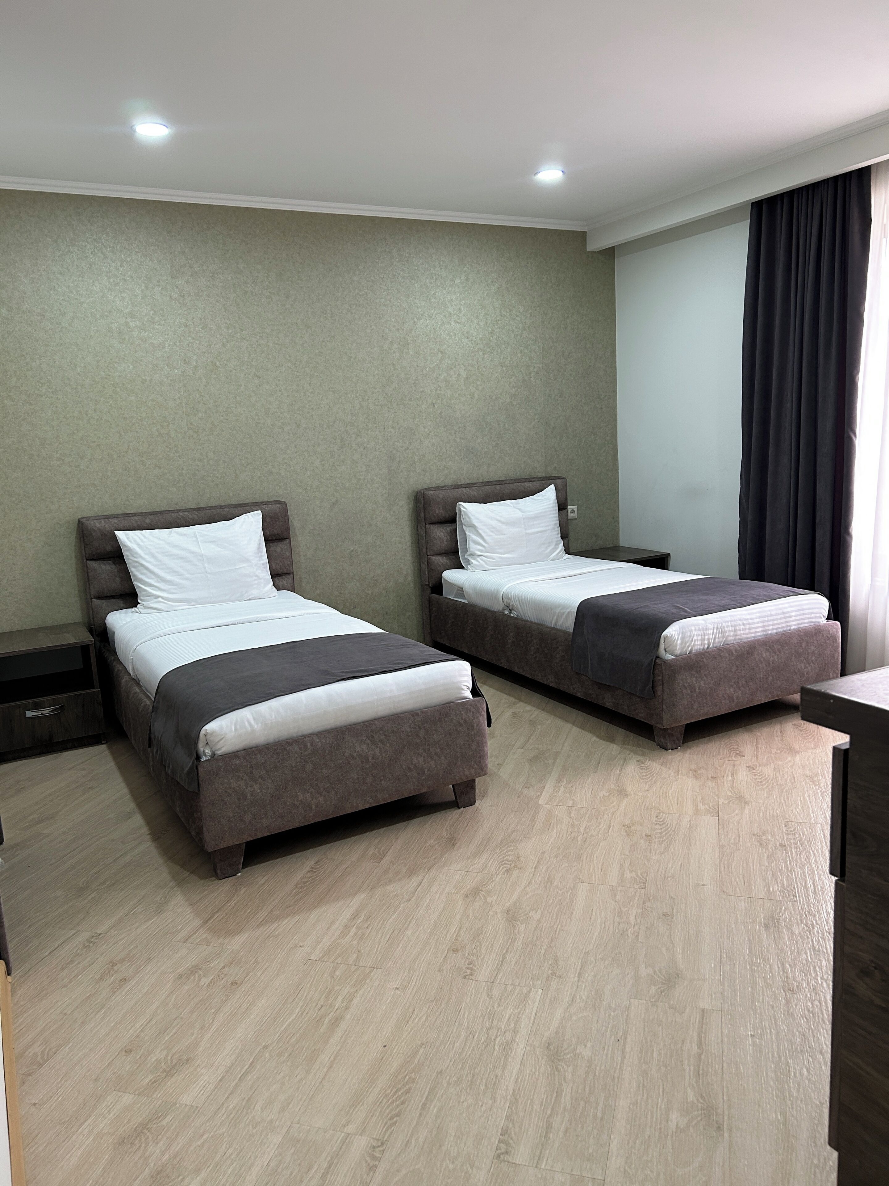 A hotel room with two beds, a wooden floor, a grey wall, and a window with curtains.