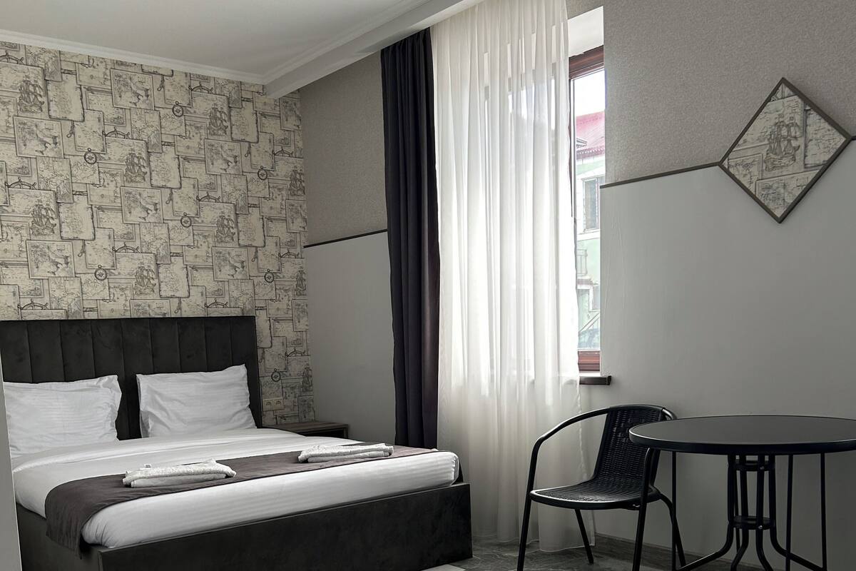 Una habitación de hotel moderna con cama, una mesita, una silla y un ventanal con cortinas.