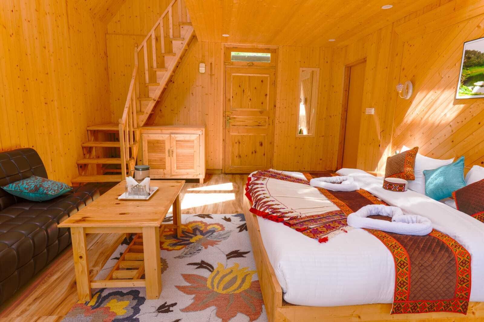 A wooden cabin interior with a staircase, a sofa, a bed with towels, and a table with a glass.