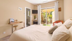 Ein Hotelzimmer mit einem Bett, einem Schreibtisch mit Stuhl, einem an der Wand befestigten Fernseher und einer Glastür, die auf einen Balkon mit Pflanzen führt.