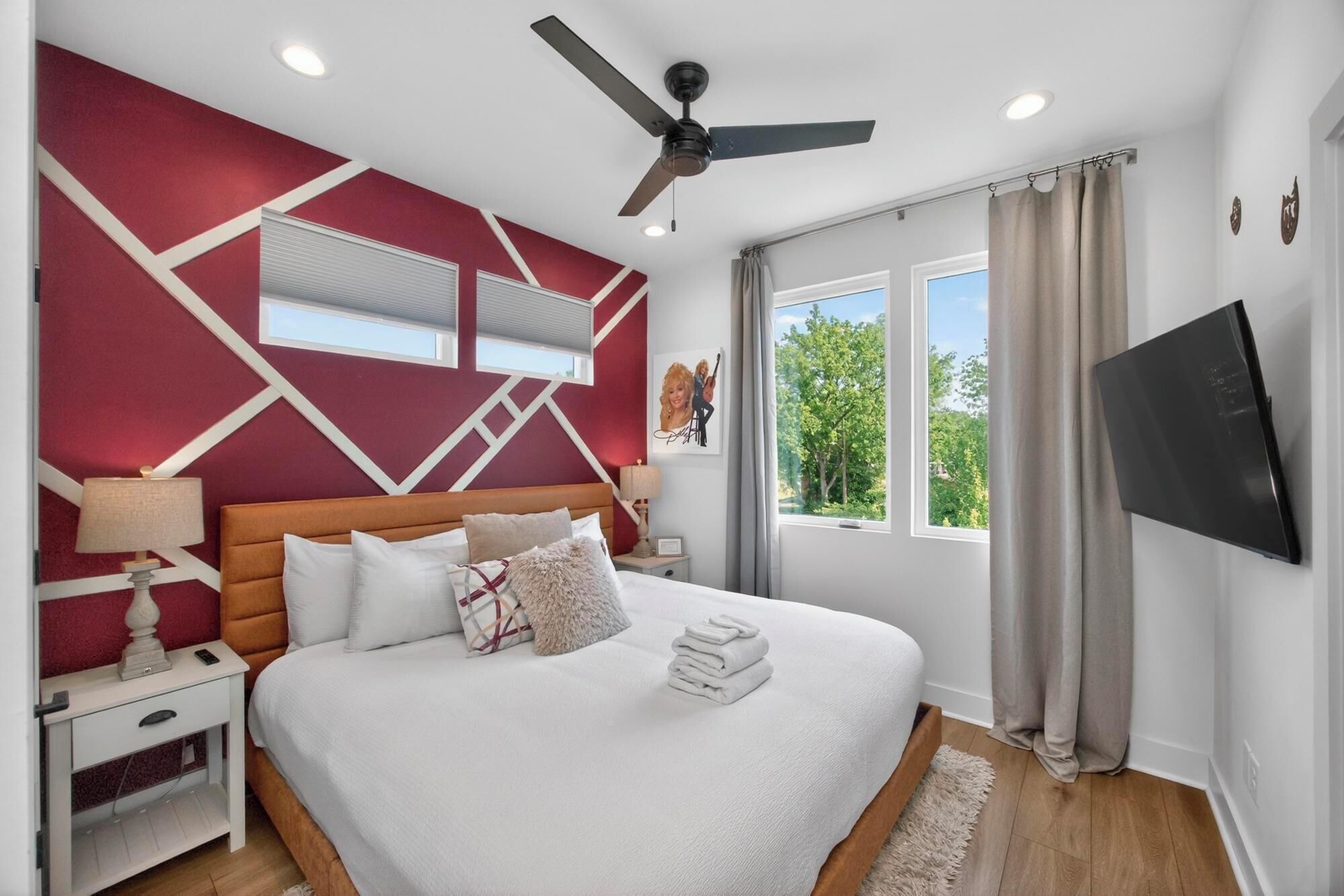 A bedroom with a bed, a nightstand, a lamp, a ceiling fan, a TV, and a window with curtains.
