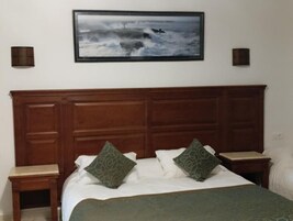Un dormitorio con cabecera de madera, dos mesitas de noche, cama con ropa blanca y almohadas estampadas de color verde, y un cuadro enmarcado de un mar embravecido en la pared.
