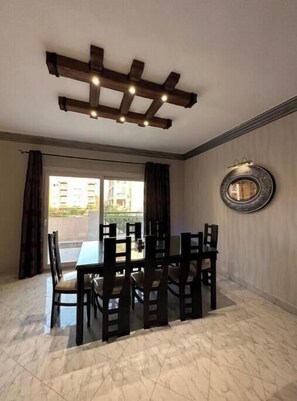 A dining area with a table and chairs, a ceiling light fixture, a window with curtains, and a wall-mounted mirror. - Pool View Villa Apartment – Private Garden Escape in Dreamland (Giza Governorate)
