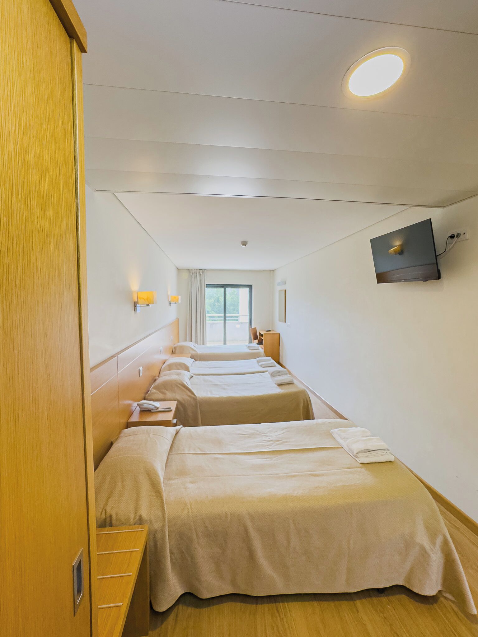 A hotel room with four beds arranged in a row, a television mounted on the wall, and a window with curtains.