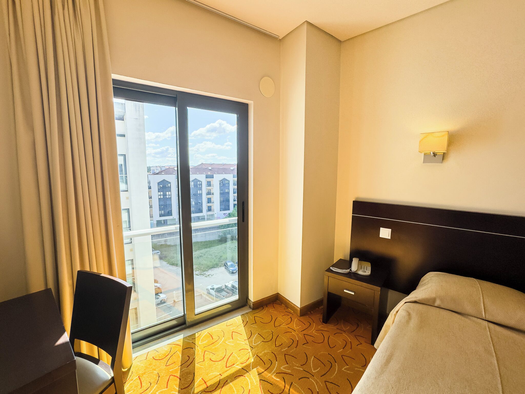 Habitación de hotel con un ventanal grande, una cama, un escritorio, una silla y una mesita de noche con lámpara.