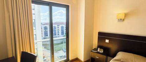 Une chambre d’hôtel avec une grande fenêtre, un lit, un bureau, une chaise et une table de chevet avec une lampe.
