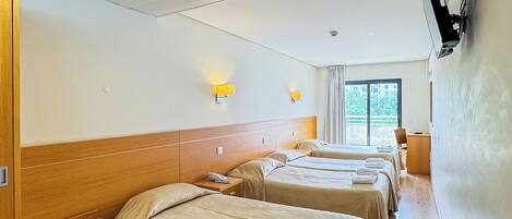 A hotel room with three beds, wooden headboards, a wooden bench, a desk, and a television mounted on the wall.
