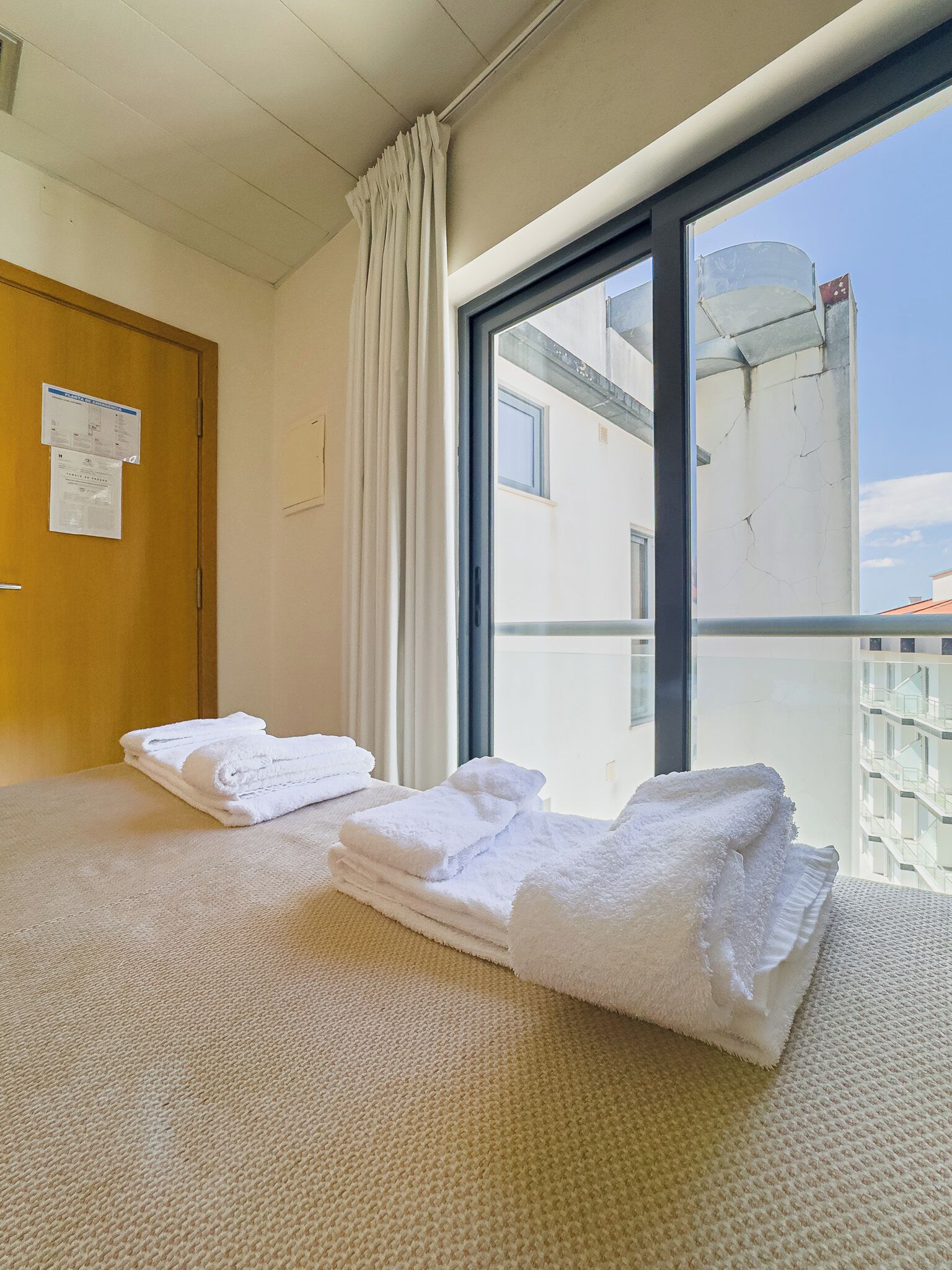 A hotel room with a large window, a bed with folded white towels, a wooden door, and a bulletin board with papers.