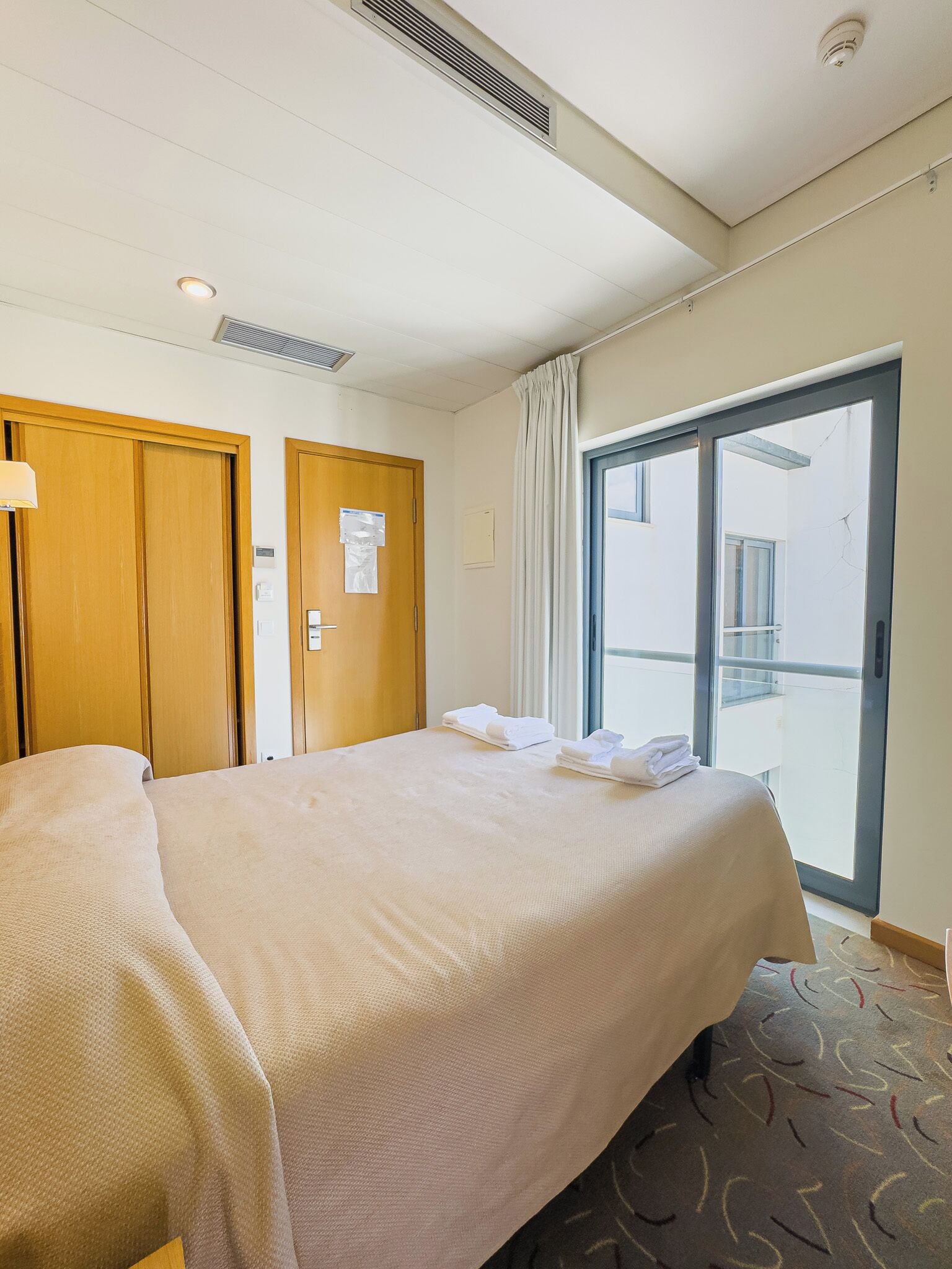 A hotel room with a bed, wooden wardrobe, a window with curtains, and a view of the outside.