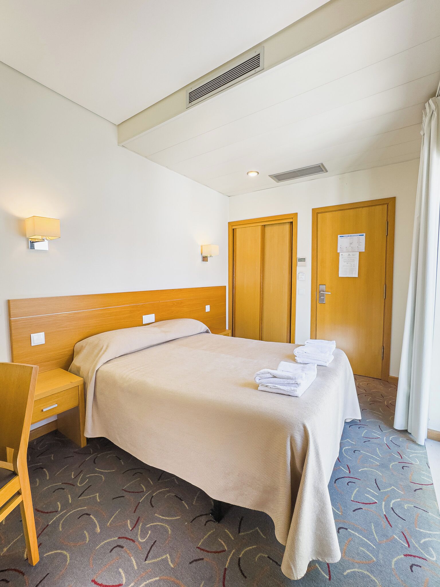 Une chambre d’hôtel avec un lit, un bureau, deux portes et une moquette à motifs.
