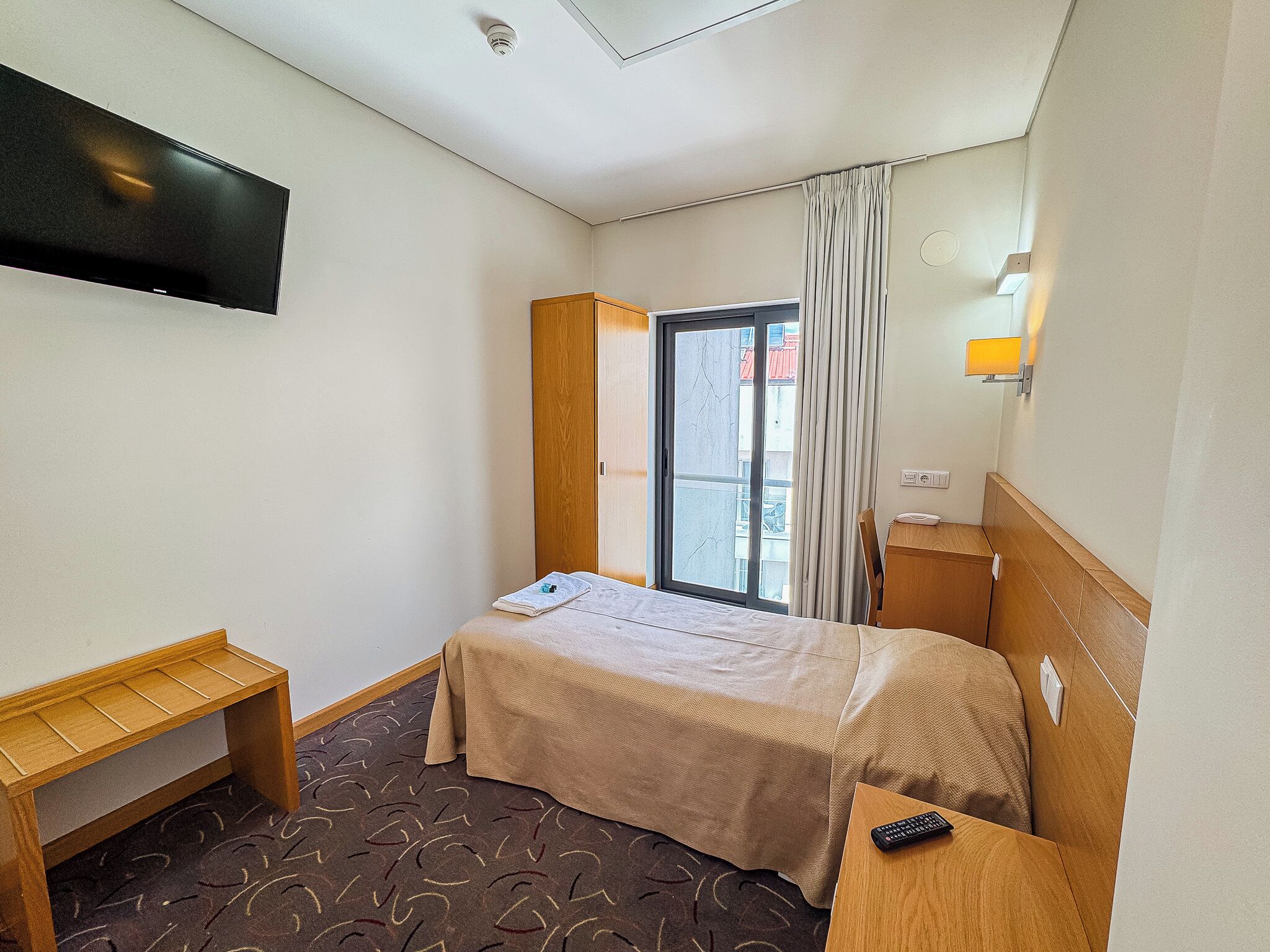 A hotel room with a bed, a wooden bench, a desk with a remote control, a window with curtains, and a wall-mounted TV.