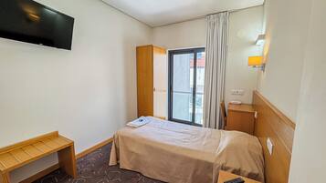 A hotel room with a bed, a wooden bench, a desk with a remote control, a window with curtains, and a wall-mounted TV.