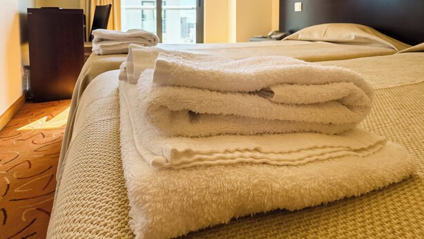 Ein Hotelzimmer mit Bett, Nachttisch, Lampe, einem Fenster mit Vorhängen und Blick nach draußen.