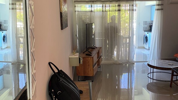Quarto de hotel moderno com um banco de madeira, televisão, escrivaninha e vista através de cortinas translúcidas.