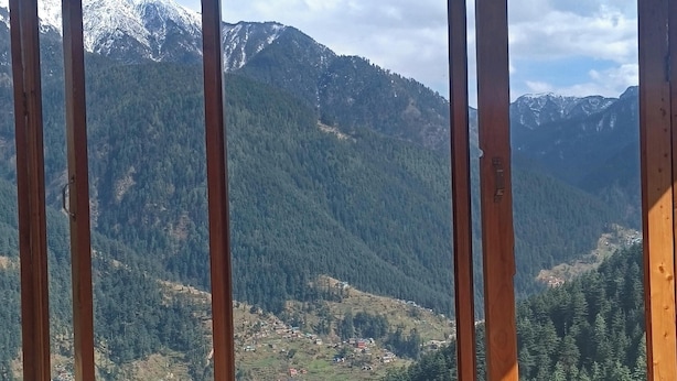 A wooden-framed window with a view of snow-capped mountains and a valley below.