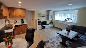 Eine moderne Küche mit Spüle, Kaffeemaschine und Schränken. Es gibt einen Essbereich mit Tisch und Stühlen sowie ein Wohnzimmer mit Sofa und Fernseher.