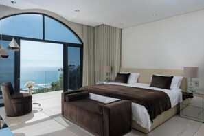 Ein modernes Schlafzimmer mit einem großen Bett, einer Couch, einem Sessel, einem Nachttisch und Blick aufs Meer.