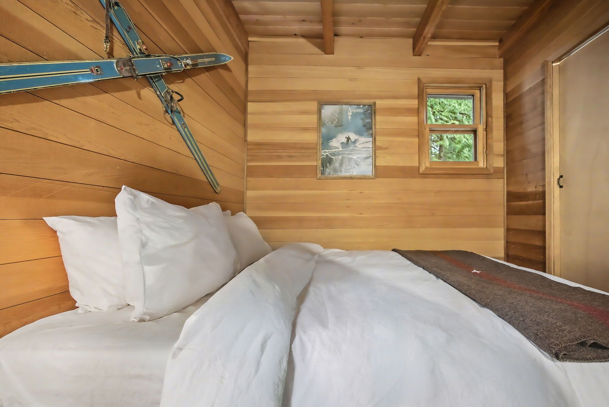 Een houten kamer met een bed, kussens, een raam en een schilderij.